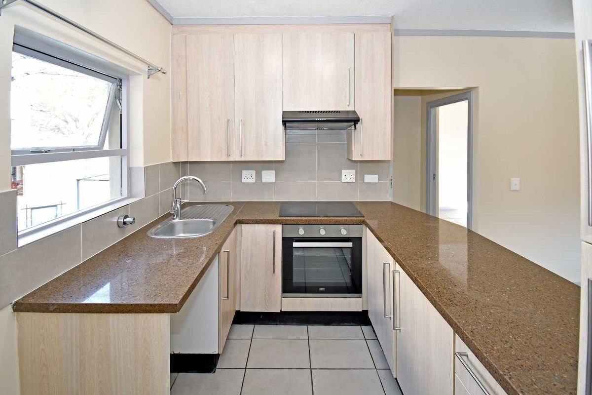 A clean, functional kitchen featuring light wood-finish cabinetry, stone countertops, integrated oven and hob, and neutral tiled flooring.
