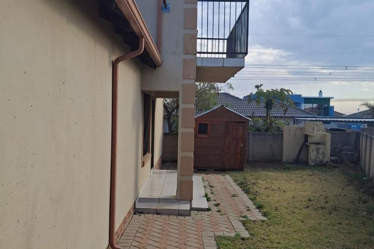 Rear exterior view of the property featuring a paved walkway, a small lawn area, a balcony with metal railing, and an outdoor braai facility.