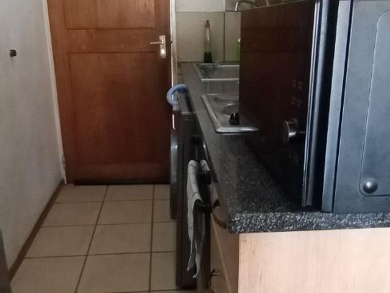 Compact galley-style kitchen featuring light wood cabinetry, dark laminate countertops, stainless steel sink, and tiled flooring.