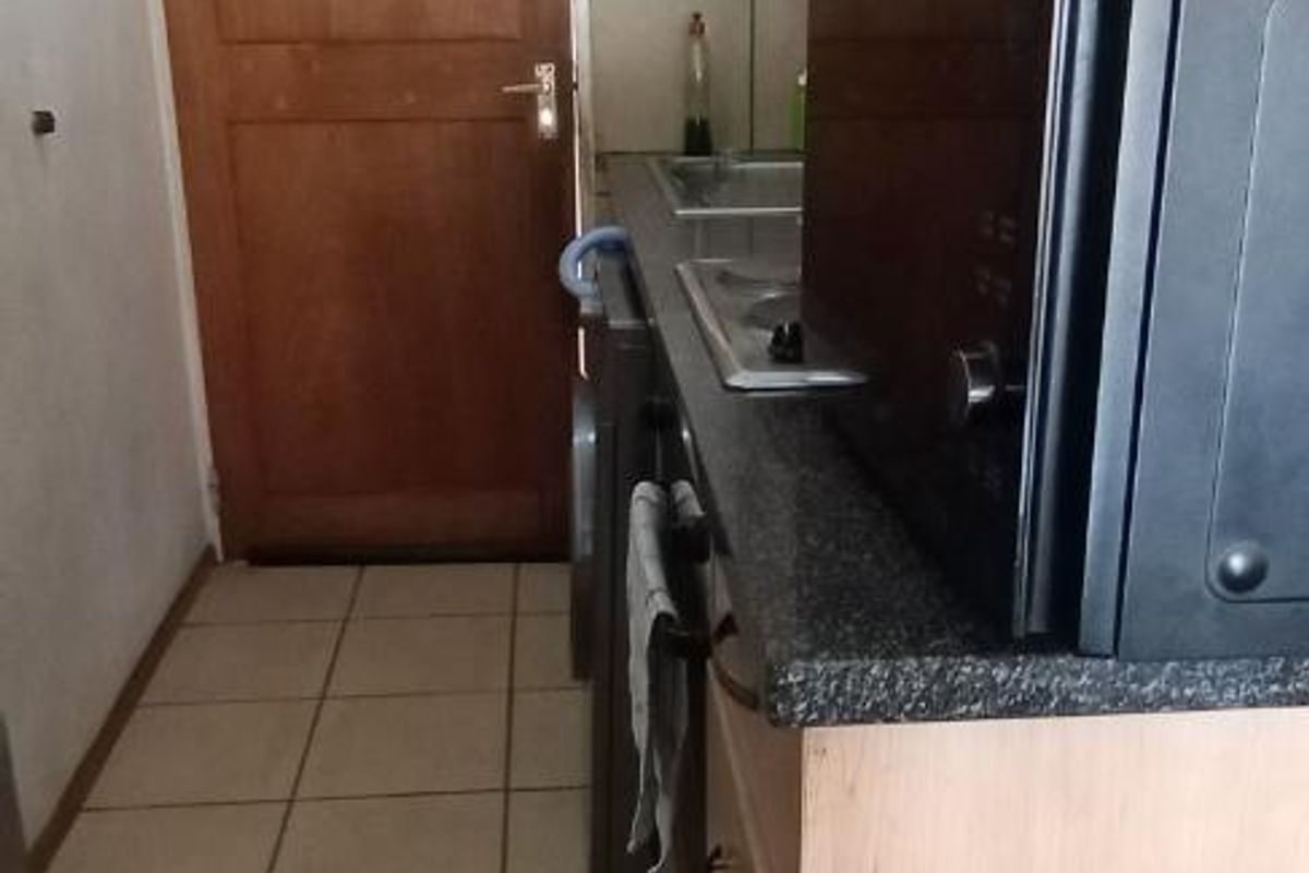 Compact galley-style kitchen featuring light wood cabinetry, dark laminate countertops, stainless steel sink, and tiled flooring.