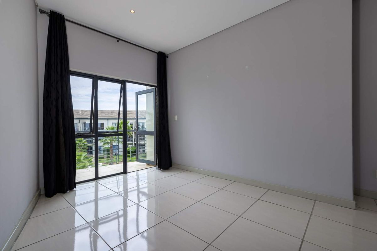 A bright, empty bedroom featuring large glass sliding doors that open onto a private balcony, finished with neutral tiled flooring.