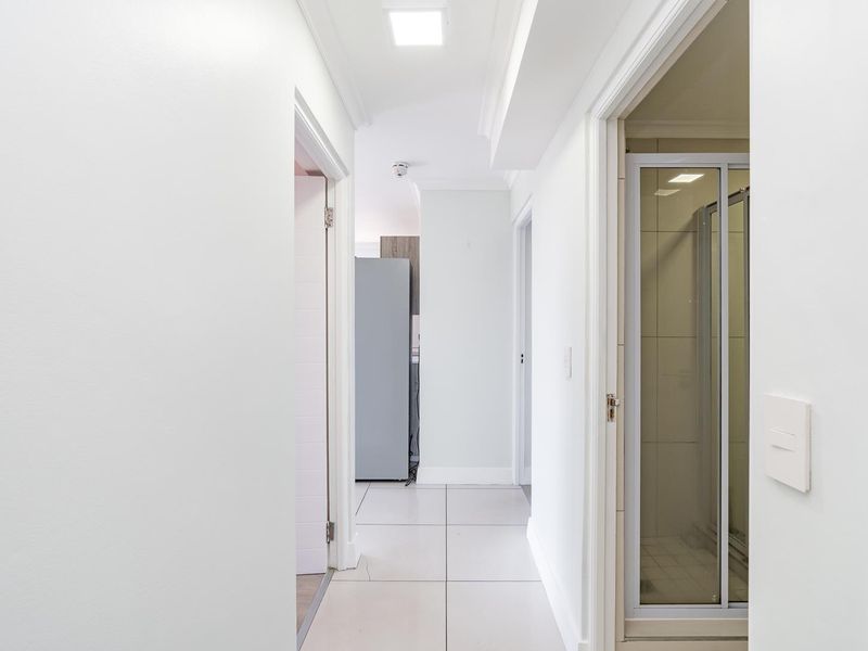A clean, well-lit hallway featuring neutral tiled flooring and recessed ceiling lights, providing access to adjacent rooms within the apartment.