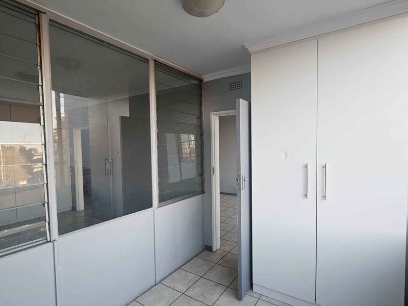 A bright, empty bedroom featuring built-in cupboards, tiled flooring, and a glass partition wall with louvered windows.