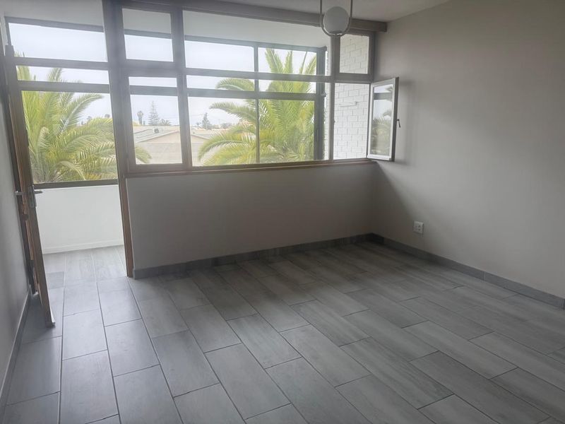 A bright, empty living room featuring modern wood-look floor tiles, large windows, and direct access to an outdoor balcony area.