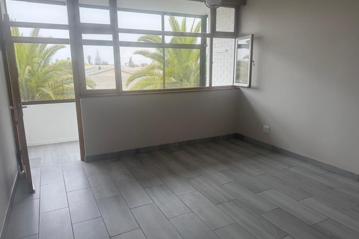 A bright, empty living room featuring modern wood-look floor tiles, large windows, and direct access to an outdoor balcony area.