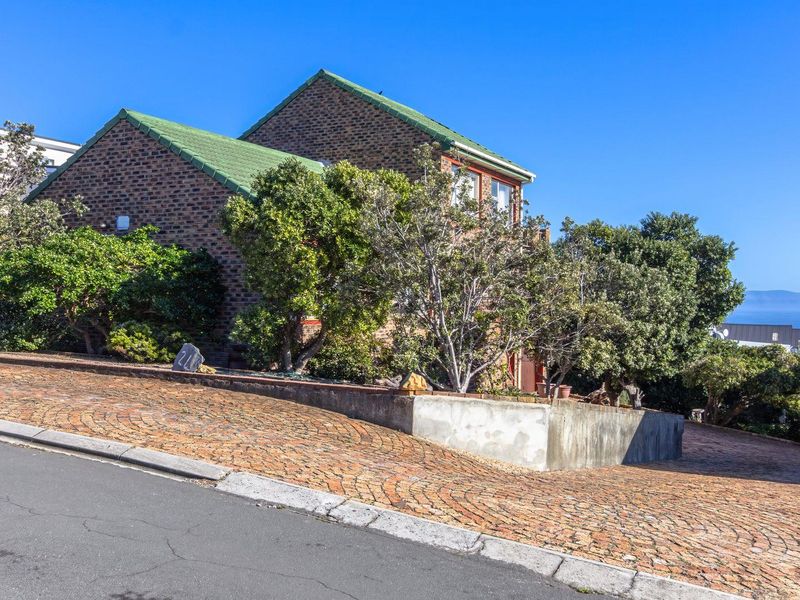 Front elevation of a brick house featuring a green tiled roof, paved driveway, and a concrete retaining wall, situated on a sloped street.