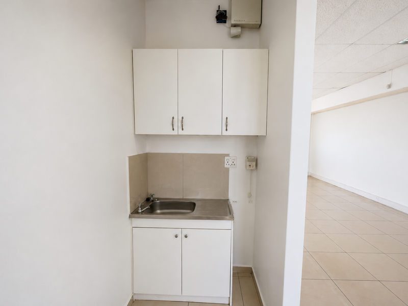 A compact kitchenette area featuring white built-in cabinetry, a stainless steel sink, and tiled splashback, adjacent to an open-plan office space.
