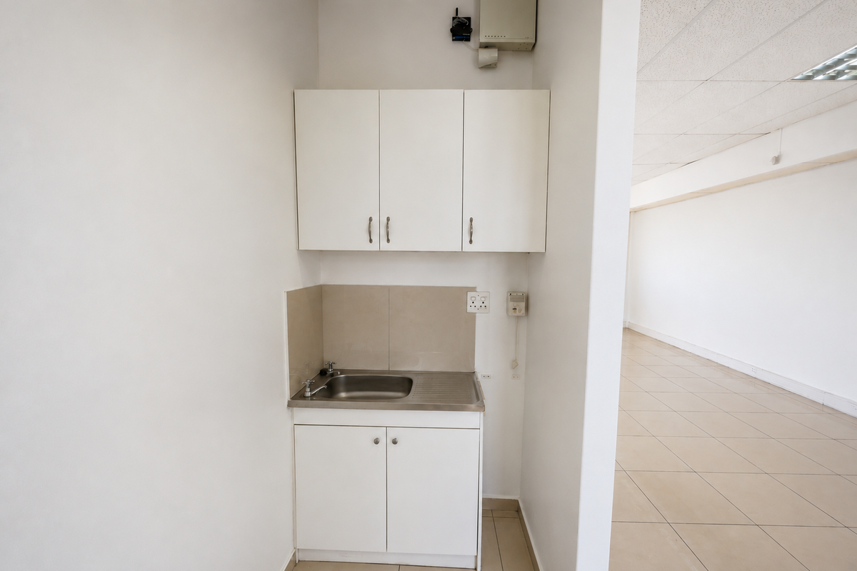 A compact kitchenette area featuring white built-in cabinetry, a stainless steel sink, and tiled splashback, adjacent to an open-plan office space.