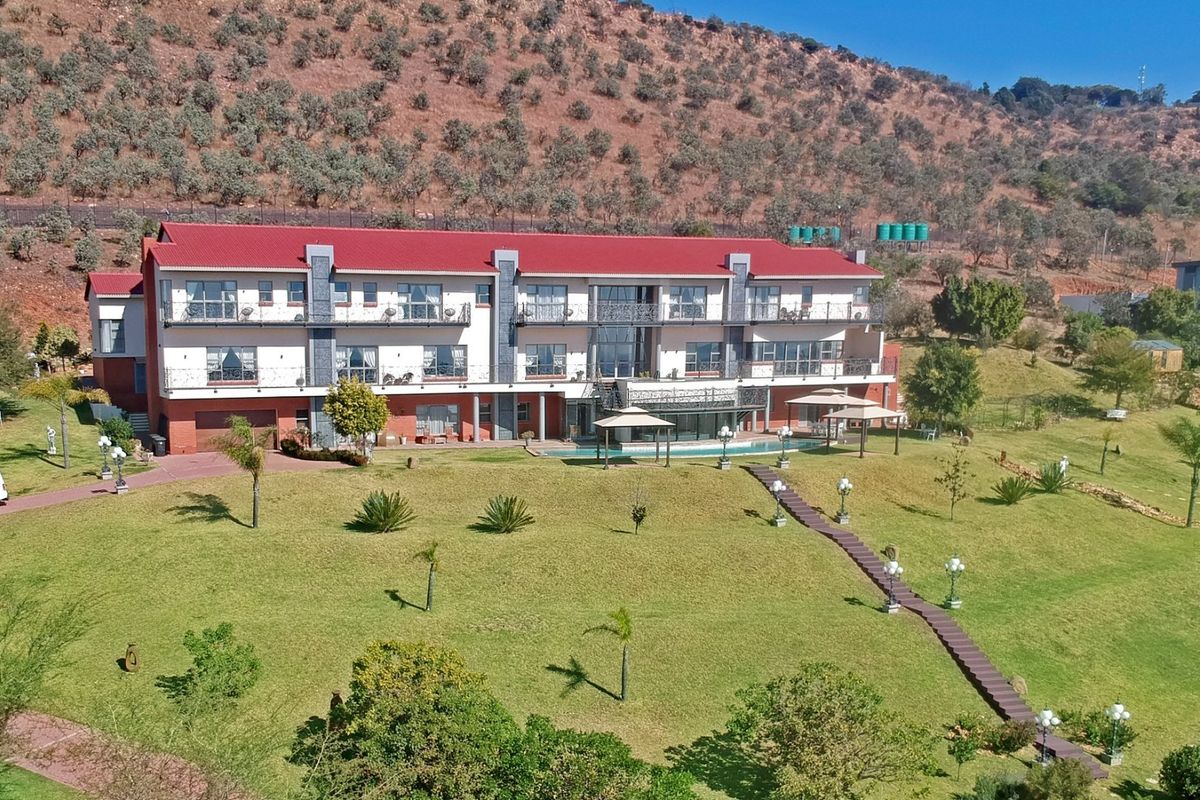 Aerial view of a large multi-story residence featuring a red roof, multiple balconies, a swimming pool, and extensive landscaped grounds set against a hillside.