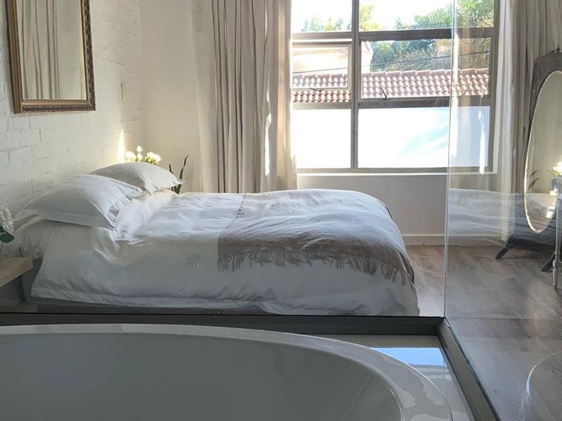 Modern bedroom featuring an open-plan design with an integrated freestanding bathtub, exposed brick wall, and large window providing ample natural light.