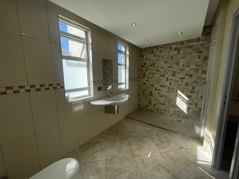 A spacious bathroom featuring a walk-in shower with mosaic tile accents, a wall-mounted basin, and neutral floor tiling with natural light.