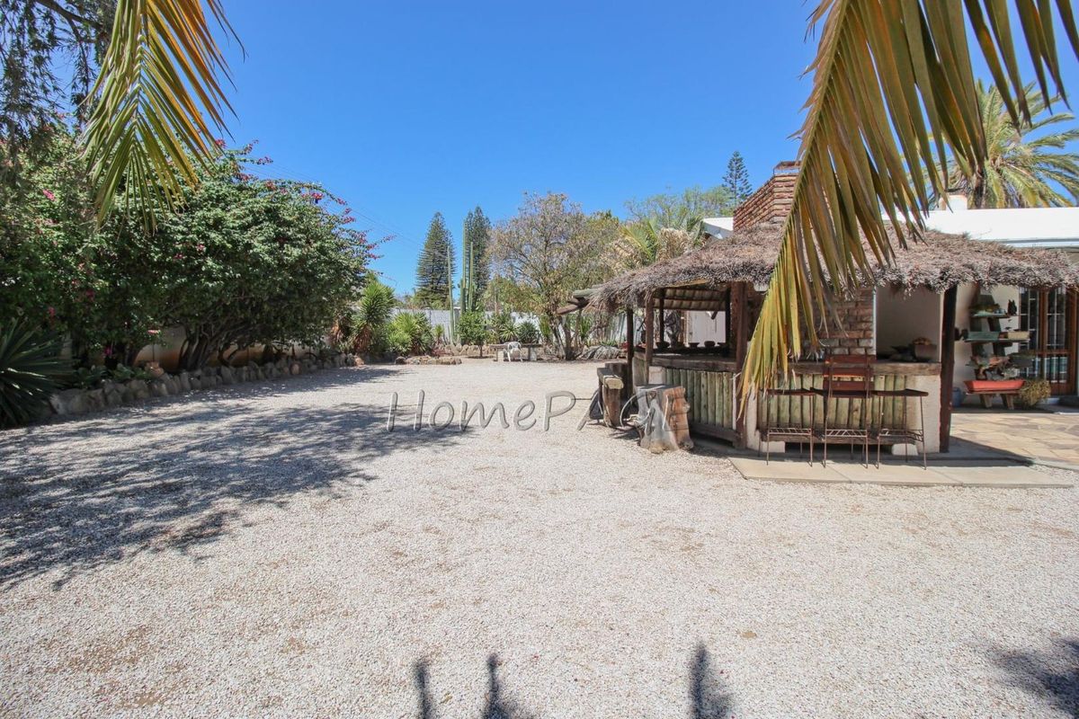 Spacious outdoor area featuring a rustic thatched-roof bar structure, gravel ground cover, and established garden surroundings.