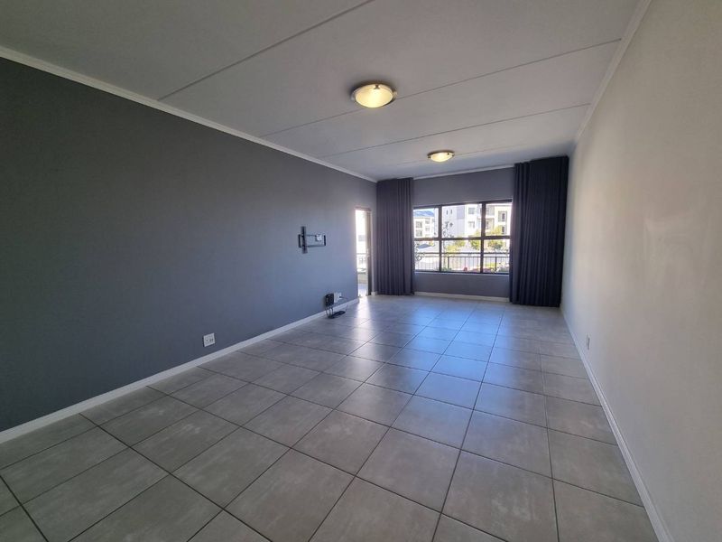 A spacious, empty living room featuring neutral grey walls, large windows with curtain tracks, and tiled flooring throughout.