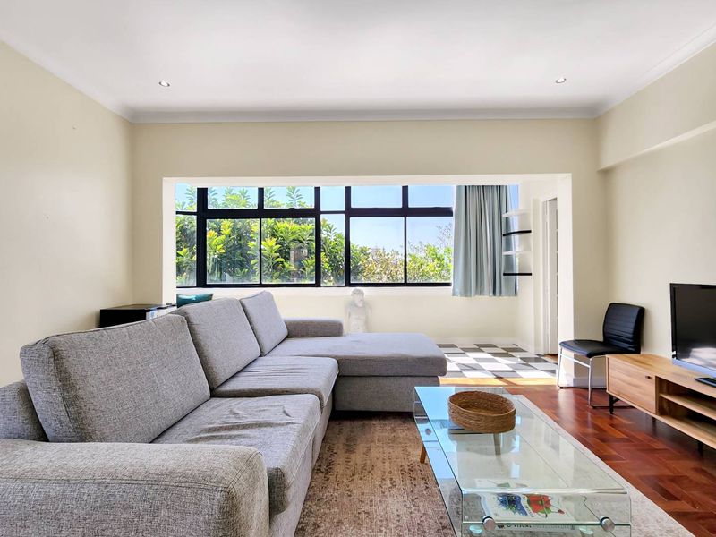 A bright living room featuring hardwood floors, recessed lighting, and a large window with views, leading to a secondary tiled area with built-in shelving.