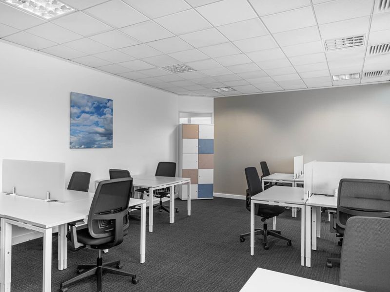 A modern, well-lit office space featuring a suspended ceiling, recessed lighting, air conditioning vents, and carpeted flooring with built-in storage.