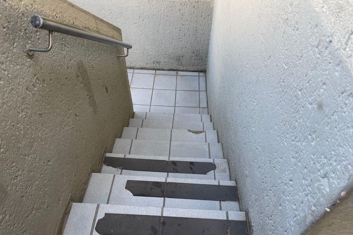 An outdoor tiled staircase featuring a stainless steel handrail and textured concrete walls, showing signs of wear on the stair treads.