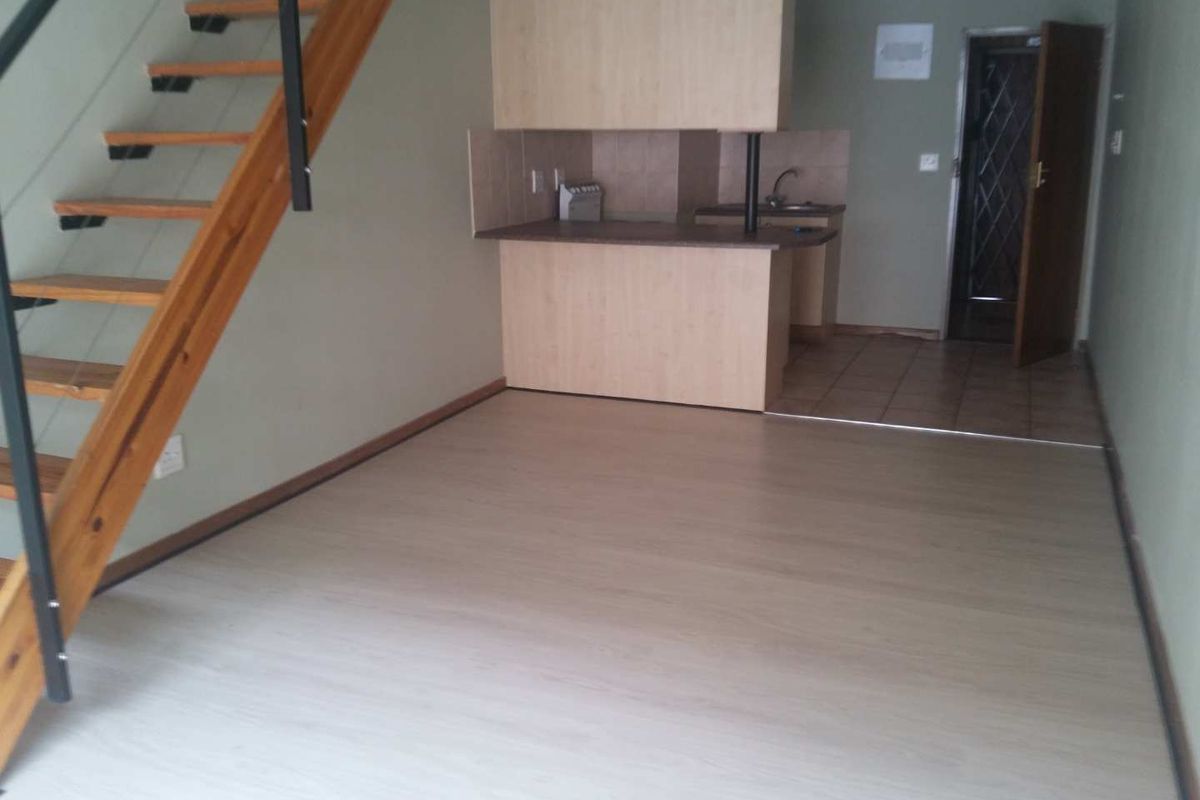 Open plan living area featuring laminate flooring, a timber staircase, and a compact kitchen with built-in cabinetry and tiled flooring.