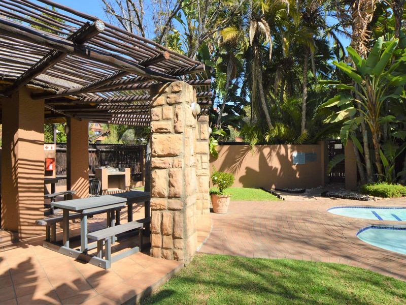 Outdoor communal pool area featuring a stone-clad pergola, built-in braai, and paved terrace surrounded by established garden landscaping.