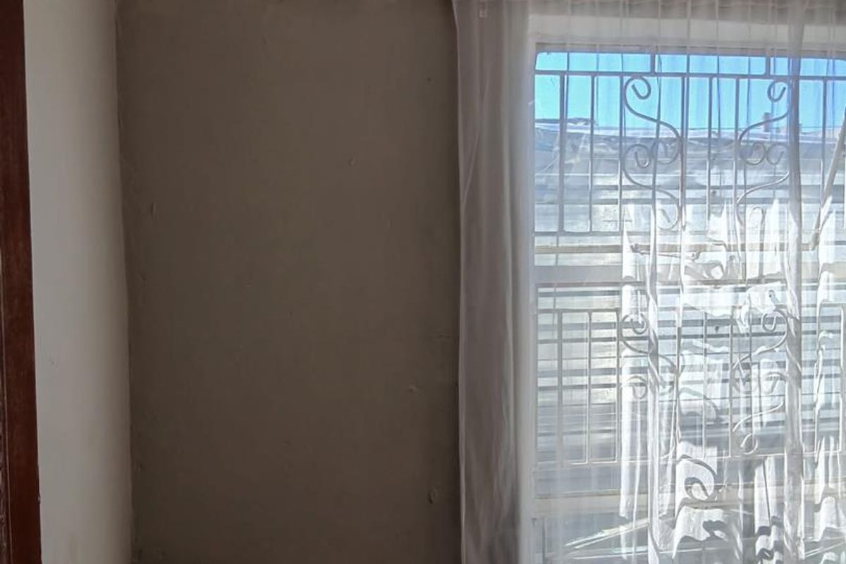 A bedroom corner featuring a window with security bars and a curtain rail, showing signs of significant wall surface deterioration.