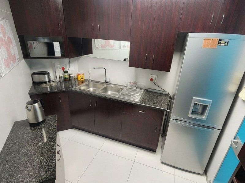 A compact office kitchen featuring dark wood cabinetry, granite countertops, a double stainless steel sink, and built-in microwave, finished with clean tiled flooring.