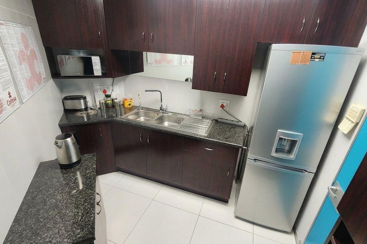 A compact office kitchen featuring dark wood cabinetry, granite countertops, a double stainless steel sink, and built-in microwave, finished with clean tiled flooring.