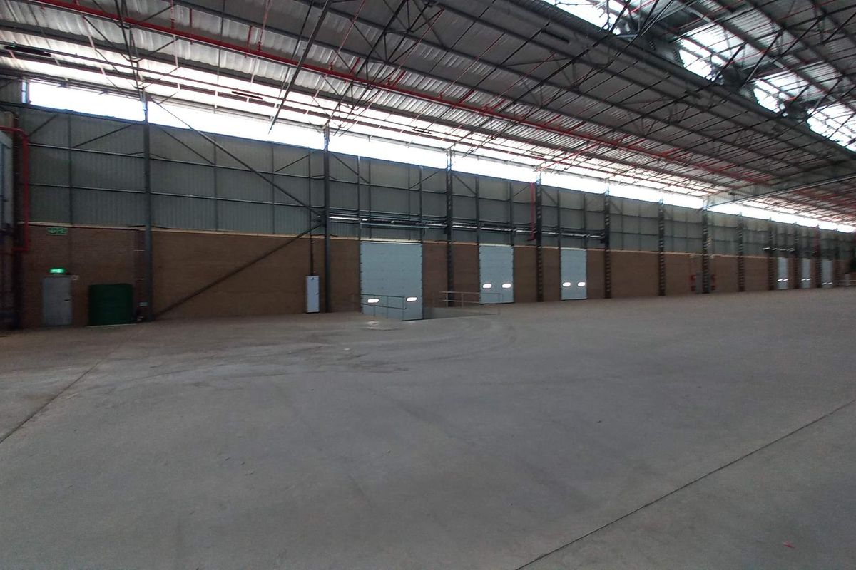 Spacious industrial warehouse featuring high ceilings with exposed steel trusses, concrete flooring, and multiple roller shutter doors for efficient loading access.
