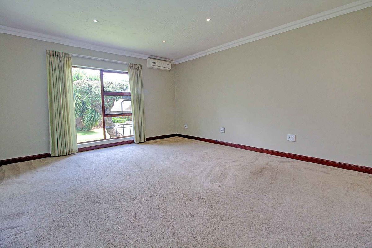 A spacious, carpeted living room featuring recessed ceiling lighting, a wall-mounted air conditioning unit, and a large window overlooking the garden.