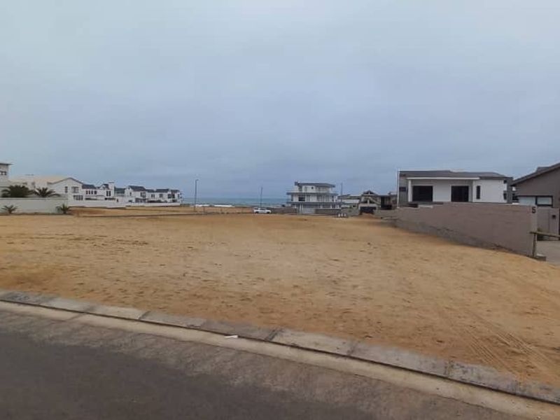 A vacant residential plot situated in a coastal neighborhood, featuring level sandy ground, boundary walls, and proximity to existing residential properties.