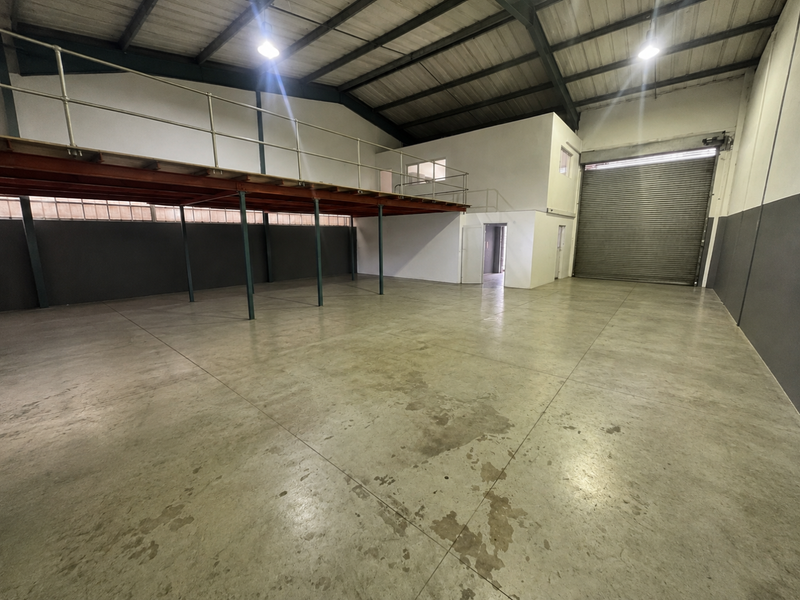 Spacious industrial warehouse unit featuring a mezzanine level, high ceilings with exposed trusses, concrete flooring, and a large roller shutter door for easy access.
