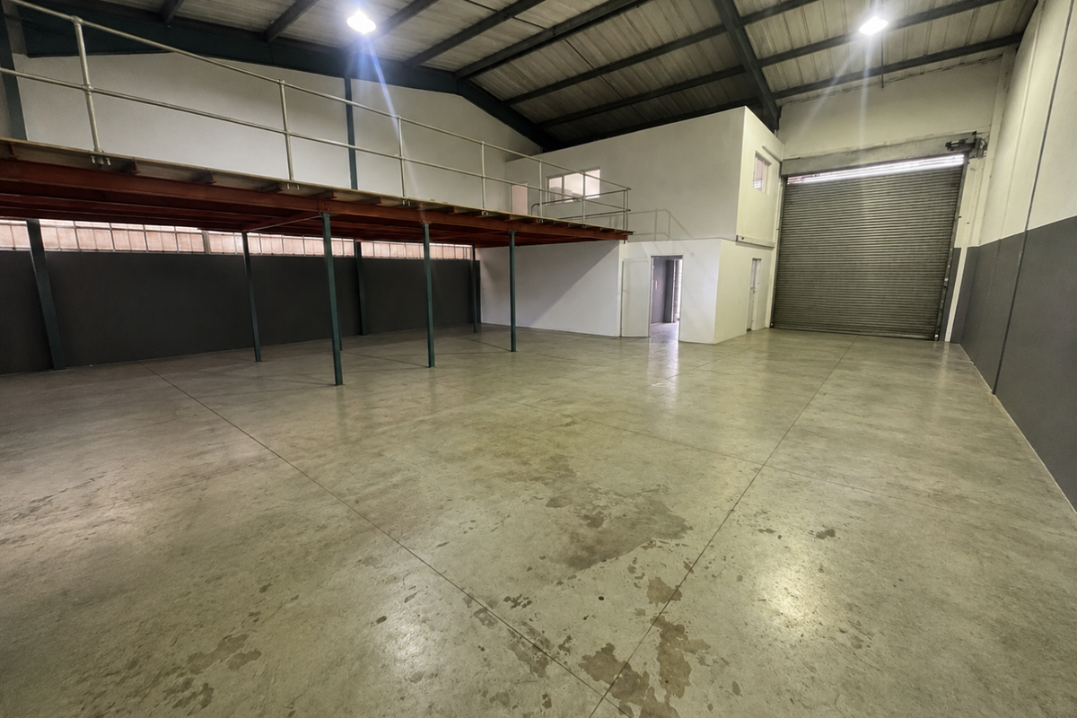 Spacious industrial warehouse unit featuring a mezzanine level, high ceilings with exposed trusses, concrete flooring, and a large roller shutter door for easy access.