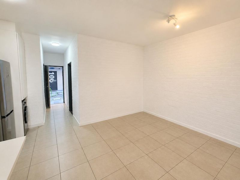 Bright open-plan living area featuring tiled flooring, white exposed brick walls, and a compact integrated kitchen space leading to the apartment entrance.
