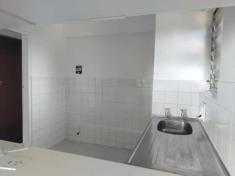 A compact, empty kitchen area featuring a stainless steel sink, tiled splashback, and louvered window, offering a blank canvas for renovation.