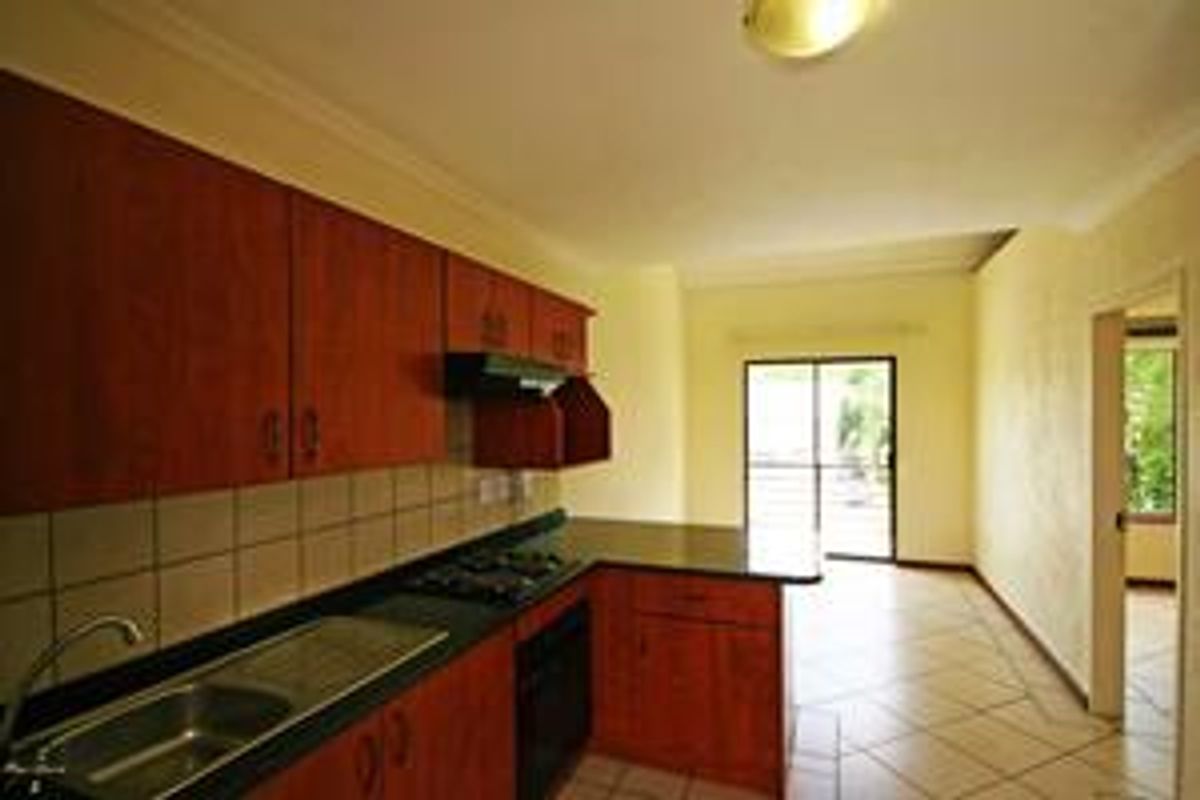 A functional kitchen featuring wood-finish cabinetry, dark countertops, built-in appliances, and tiled flooring, leading to an adjacent living area with sliding door access.