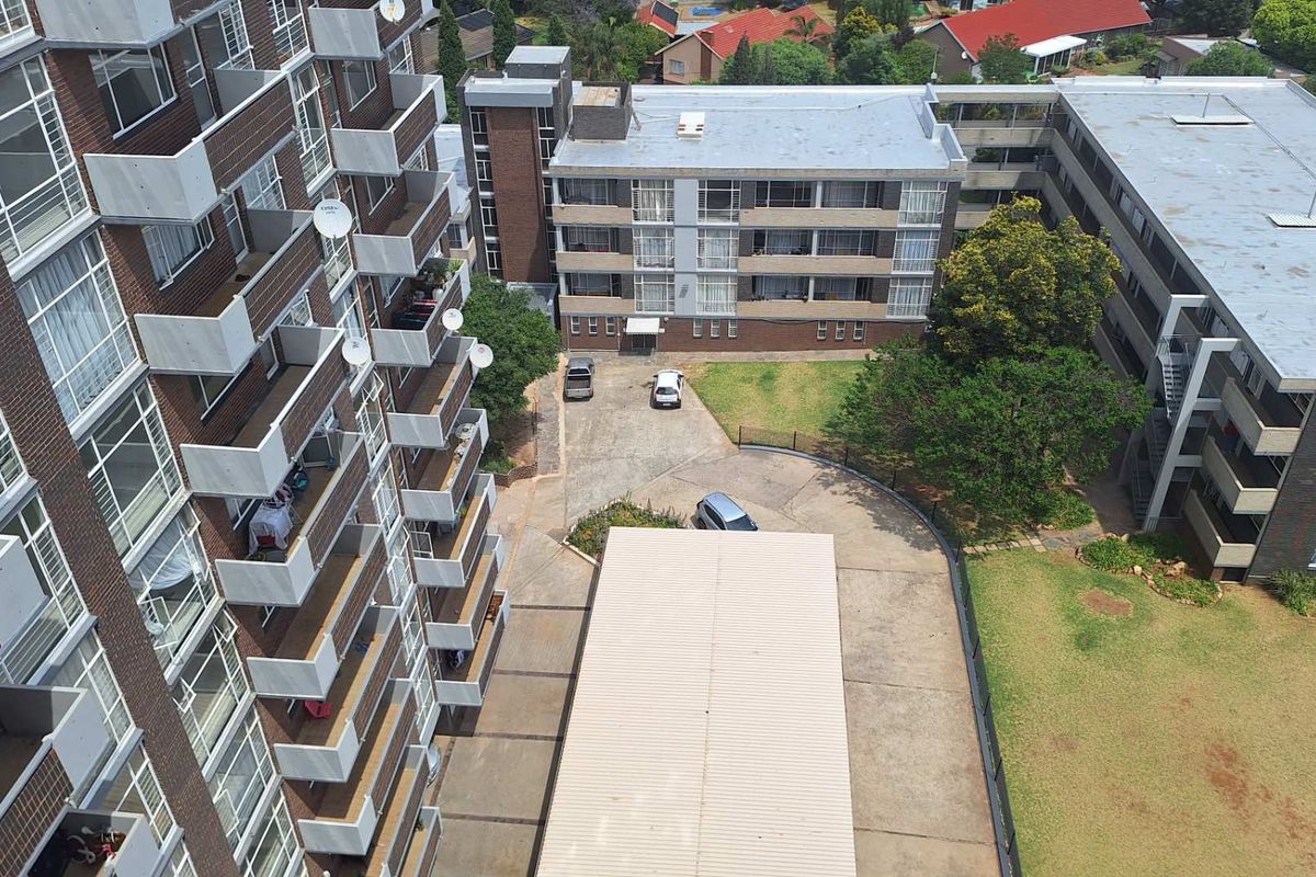 An elevated aerial view of an apartment complex featuring brick facades, multiple balconies, a paved driveway, covered parking, and communal garden areas.