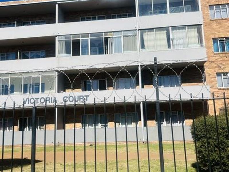Front elevation of Victoria Court apartment building featuring brick facade, enclosed balconies, and secure perimeter fencing with razor wire.