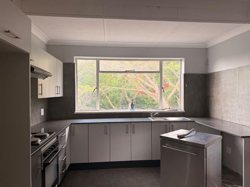 A functional kitchen featuring granite countertops, built-in cabinetry, a stainless steel sink, and an integrated oven with extractor fan, overlooking a leafy garden view.