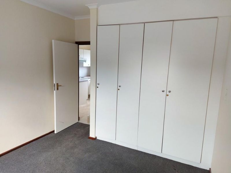 A clean, empty bedroom featuring neutral walls, grey carpeting, and ample built-in cupboard storage, with a doorway leading to an adjacent kitchen area.