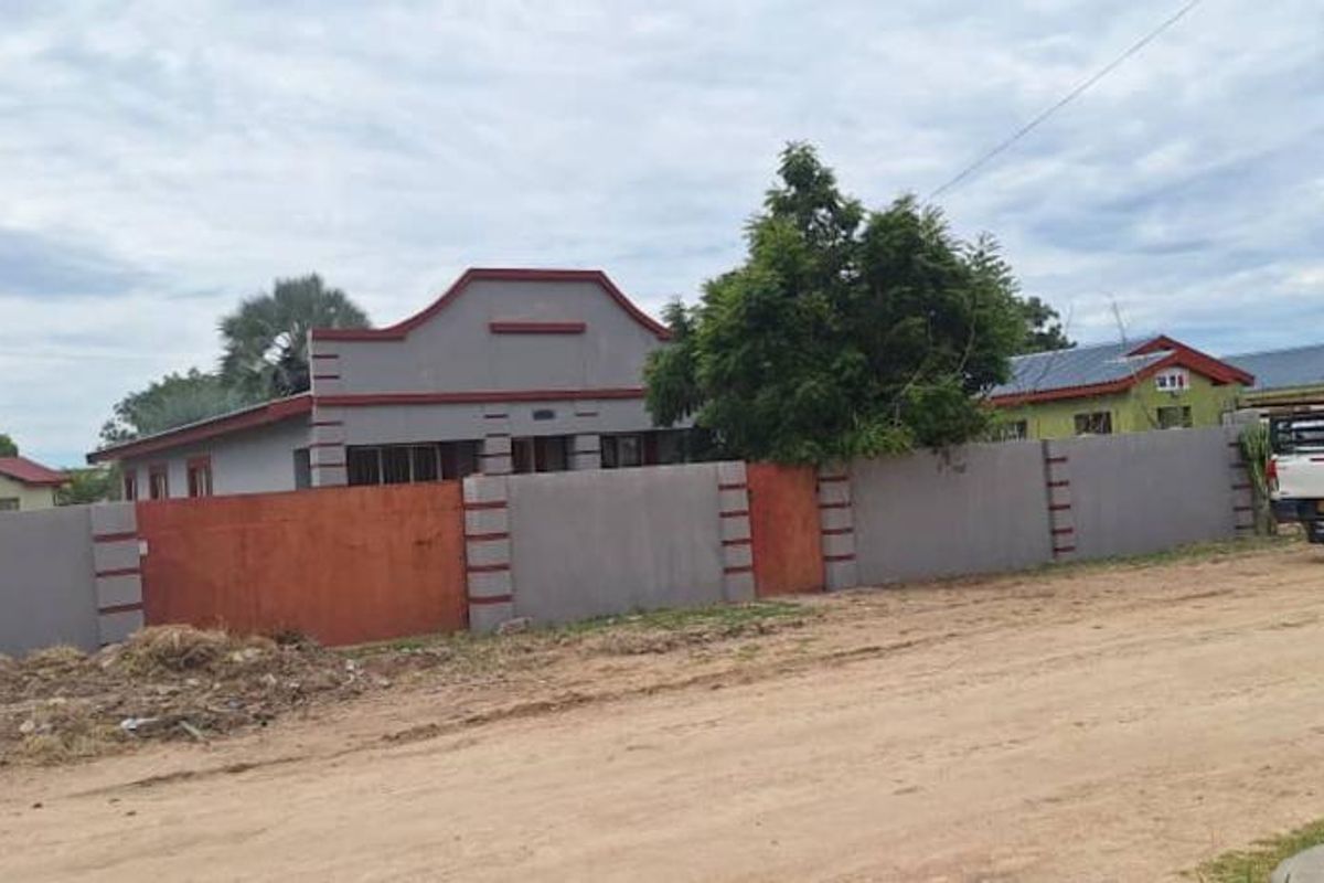 Front view of a residential property featuring a perimeter wall and metal roof, situated along an unpaved road.