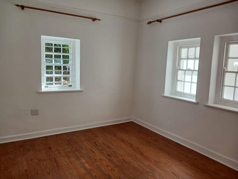 A bright, empty bedroom featuring polished timber flooring, classic sash windows with security bars, and neutral white walls.