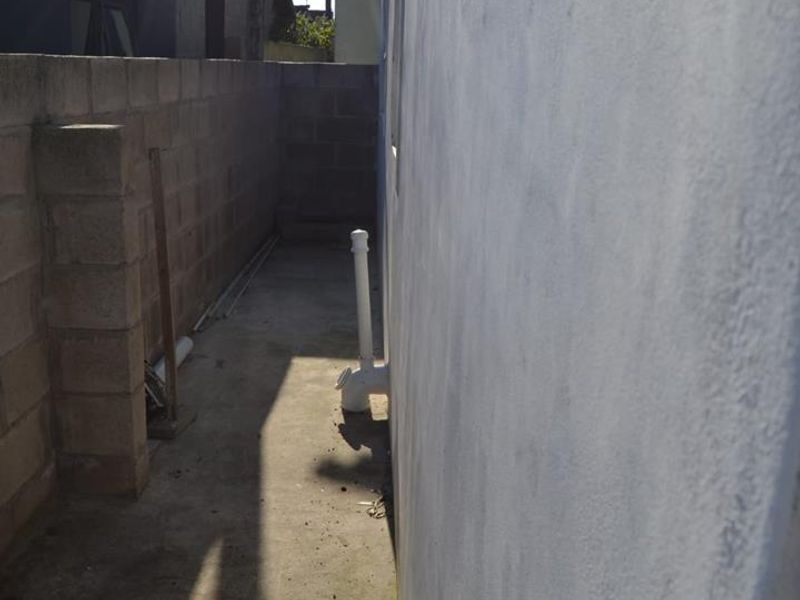 A narrow side passage featuring a concrete pathway and boundary wall with exposed external plumbing infrastructure.