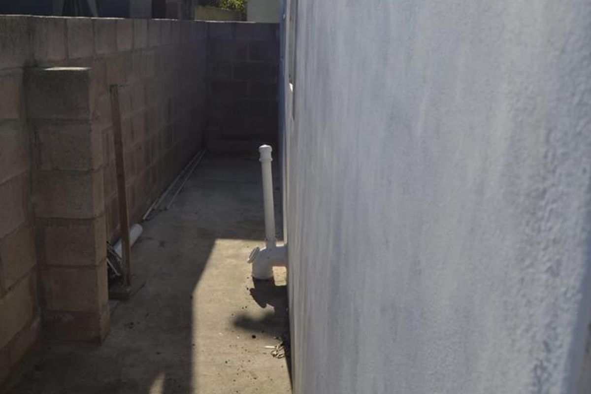 A narrow side passage featuring a concrete pathway and boundary wall with exposed external plumbing infrastructure.