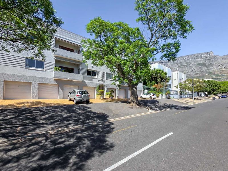 Street-level view of a multi-story apartment building featuring multiple garage doors, balconies, and mature landscaping with mountain views in the background.