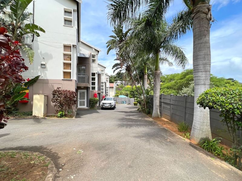 A view of the complex driveway and front facade, featuring paved access, boundary walling, and established palm trees.