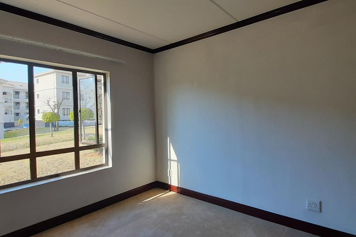 A bright, empty bedroom featuring neutral walls, dark skirting boards, and a large aluminium-framed window overlooking the complex grounds.