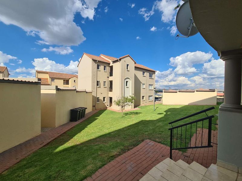 A view of the apartment complex rear exterior, featuring a communal lawn, brick paved pathways, and boundary walls.