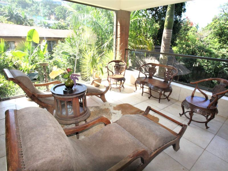 A spacious covered balcony featuring tiled flooring, a modern glass balustrade, and a stone pillar, offering a pleasant outdoor seating area with garden views.