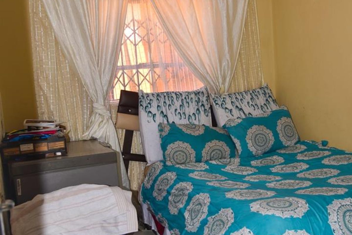 A bedroom featuring tiled flooring and a window with security bars, showing signs of wear including ceiling water staining and wall paint deterioration.