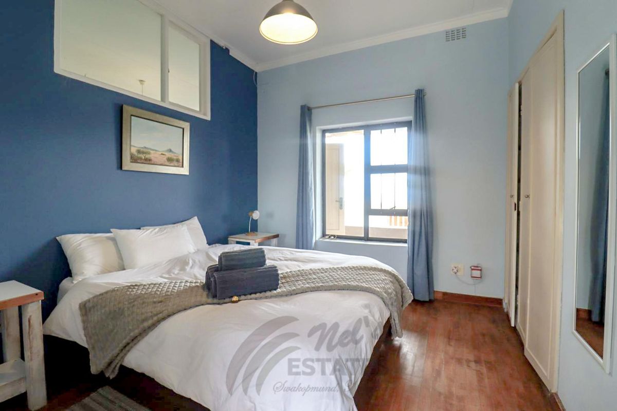 A bedroom featuring timber flooring, built-in cupboards, and a window allowing natural light, finished with a blue accent wall.