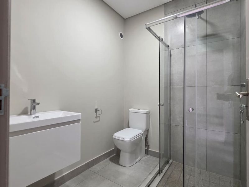 A modern, clean bathroom featuring a wall-mounted vanity, a toilet, and a glass-enclosed shower with grey tiled finishes.