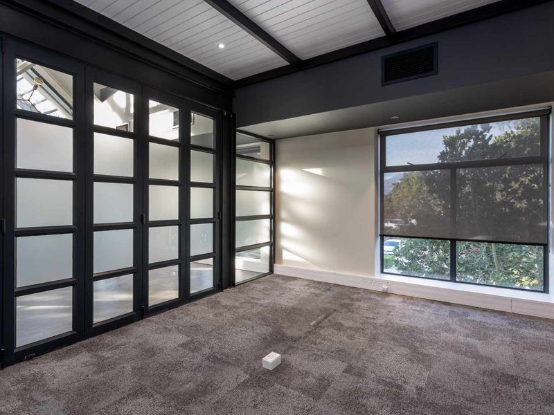 A modern, empty office space featuring dark-framed glass partitions, carpeted flooring, and large windows that provide ample natural light.
